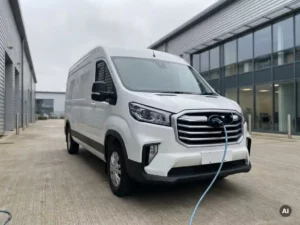 Witte elektrische Maxus bezorgbus op bedrijventerrein met laadkabel, moderne magazijnfaçade op de achtergrond.