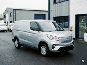 Maxus elektrische bedrijfsbus geparkeerd op een dealerterrein met industrieel magazijn op de achtergrond, driekwart vooraanzicht.